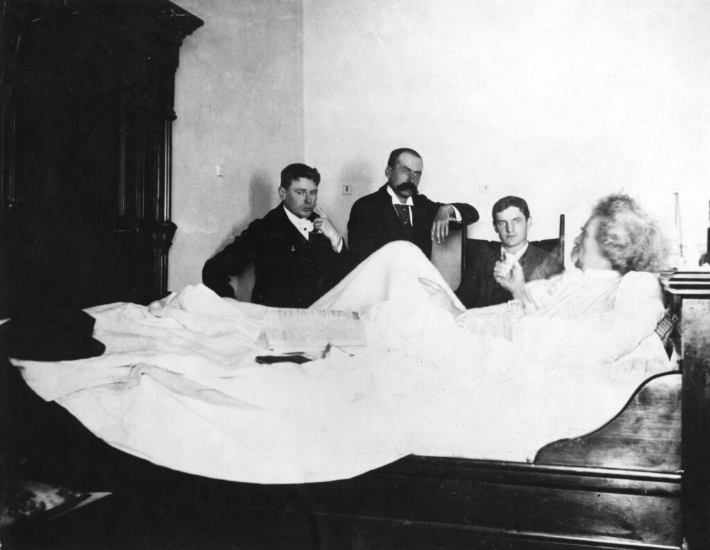 Mark Twain sitting in a bed speaking to three male reporters sitting around him listening.